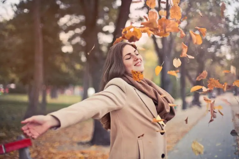 Young woman in a beige coat joyfully throwing autumn leaves in a park.
